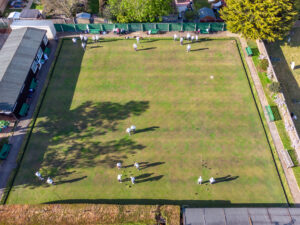 Drone shot of the outdoor green from above on a sunny day, with players scattered across the rinks and trees visible at the boundary