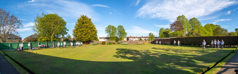 Panoramic view of the outdoor bowling green at Swanscombe Park on a sunny day, with members playing and trees in the background